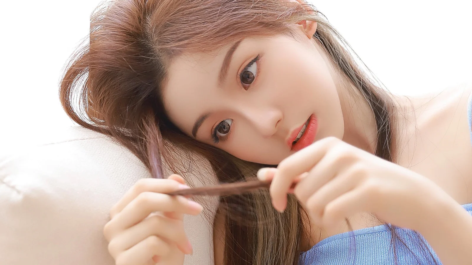 긴 머리의 여성이 밝은 배경 앞에 누워 파란색 상의를 입고 머리카락 한 가닥을 손에 들고 있습니다.