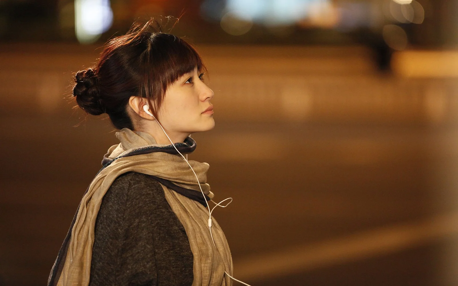 一名戴着耳机、围着围巾的女性在暖色虚化背景前侧身仰望。