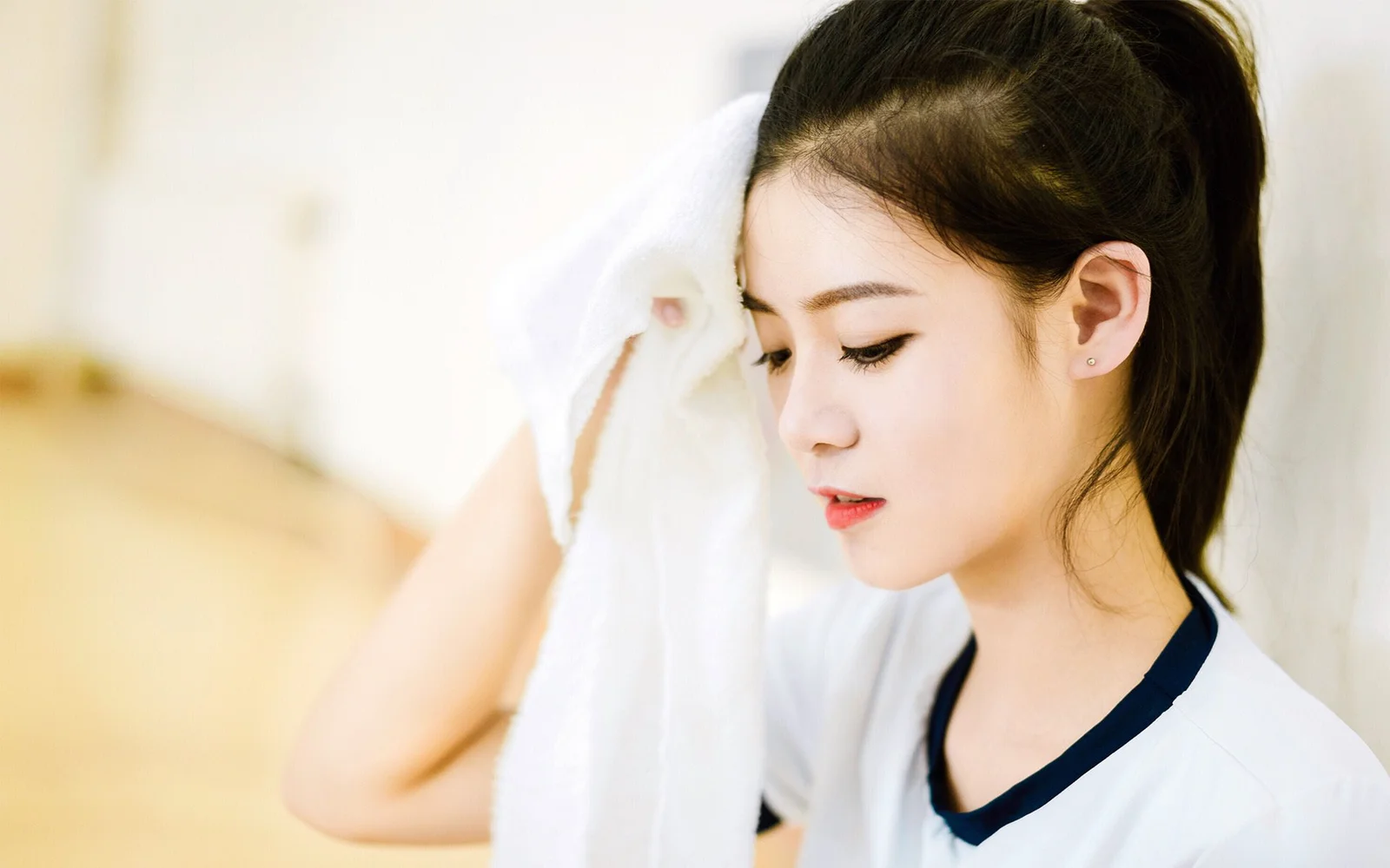 ポニーテールの女性が白いトップスを着て、明るい室内で白いタオルを持ちながら横向きに立っています。
