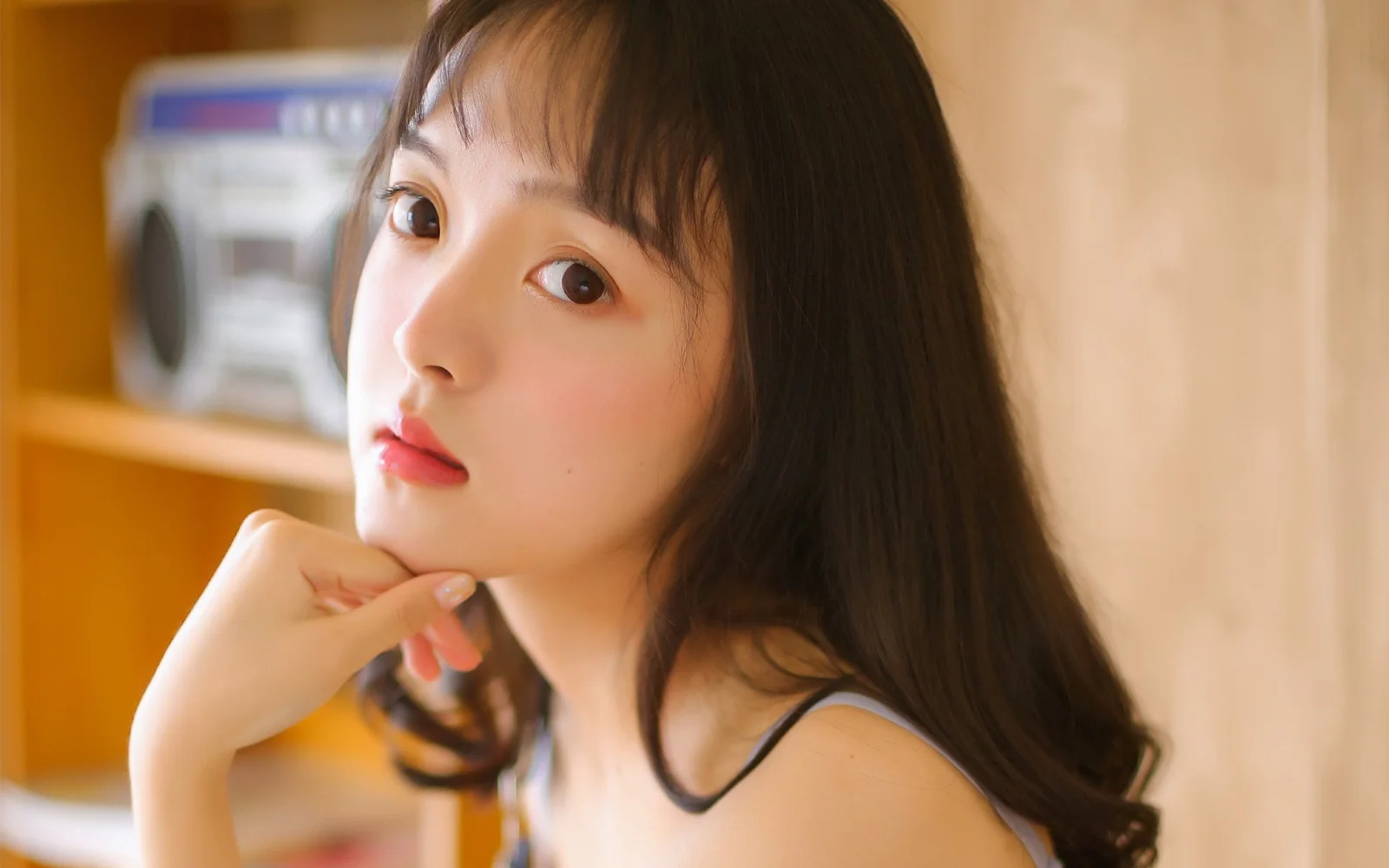 一位長髮女性在暖色室內背景前托腮注視鏡頭。