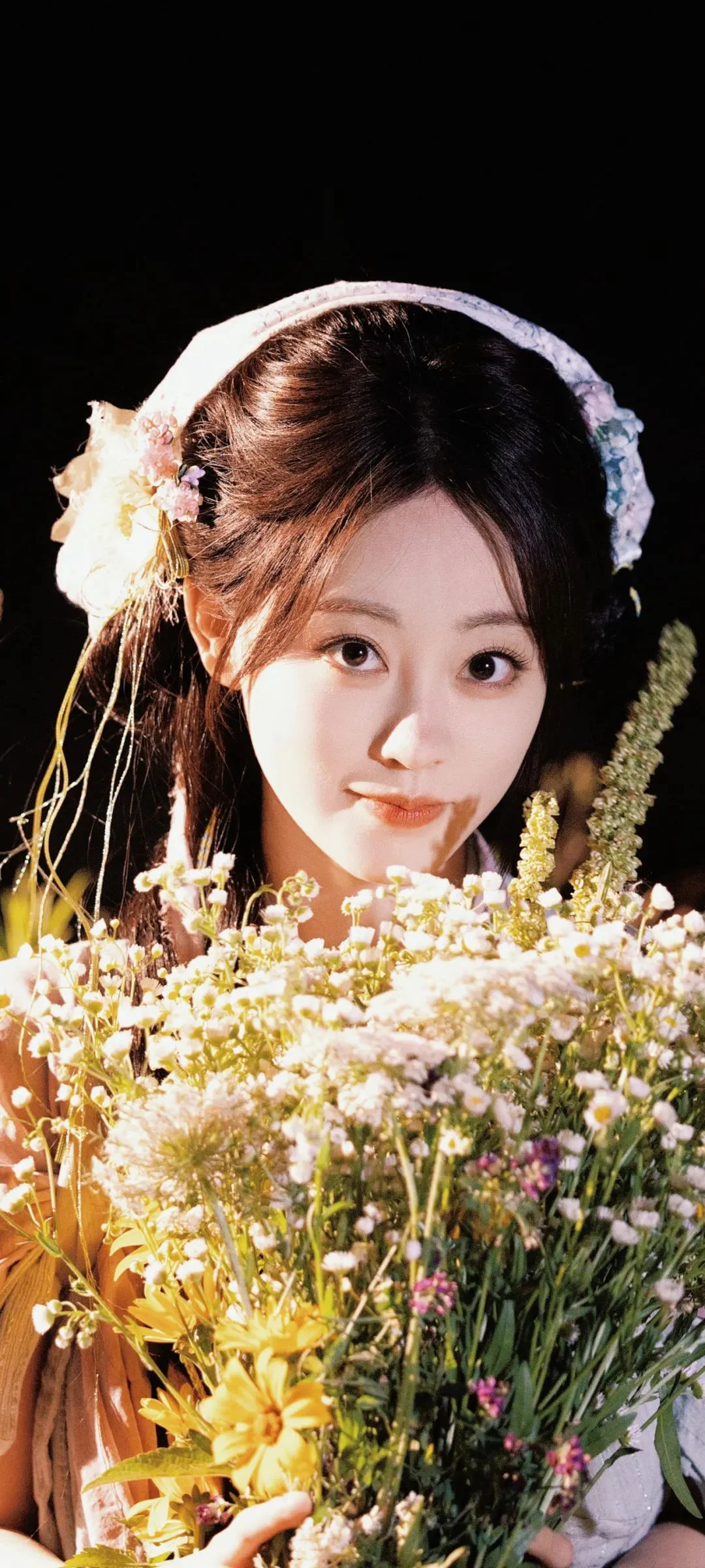 검은 배경 앞에서 성인 여성이 꽃다발을 들고 흰색과 노란색 꽃들에 둘러싸여 있다.