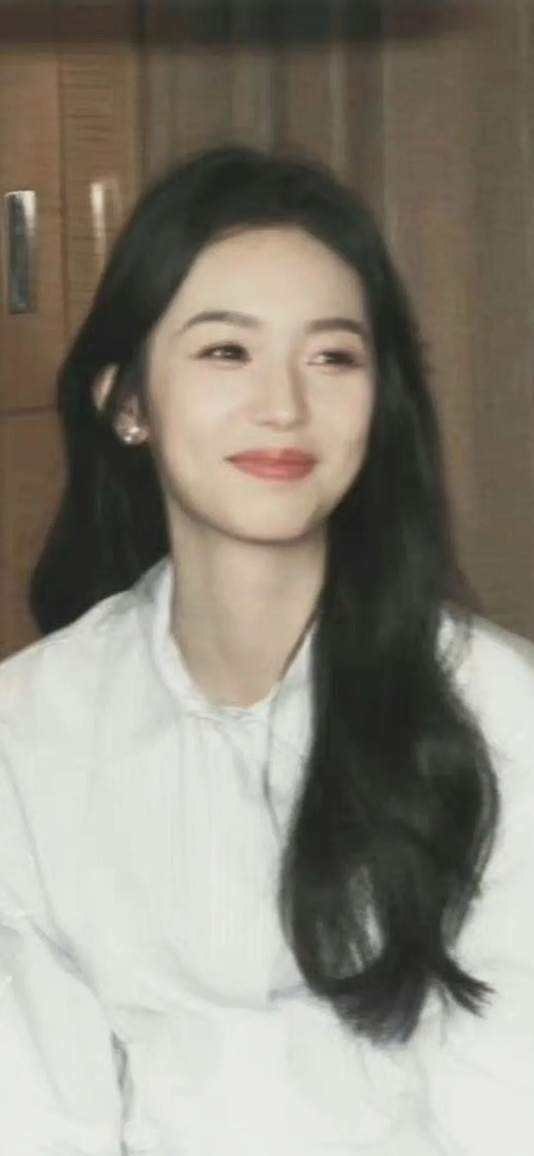 一名长发女性身穿白色上衣，在室内木质背景前微笑看向一侧。