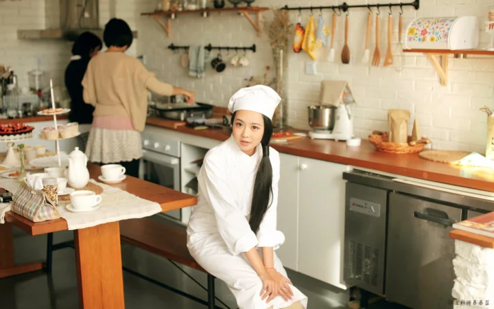 一名穿白色厨师服和帽子的女性坐在明亮的厨房中。