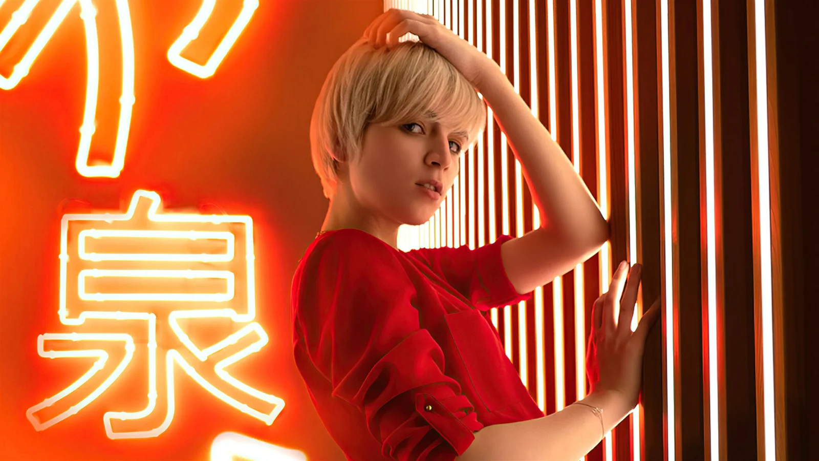 一名穿红色上衣的金发女子站在明亮的霓虹灯和竖直光带前。