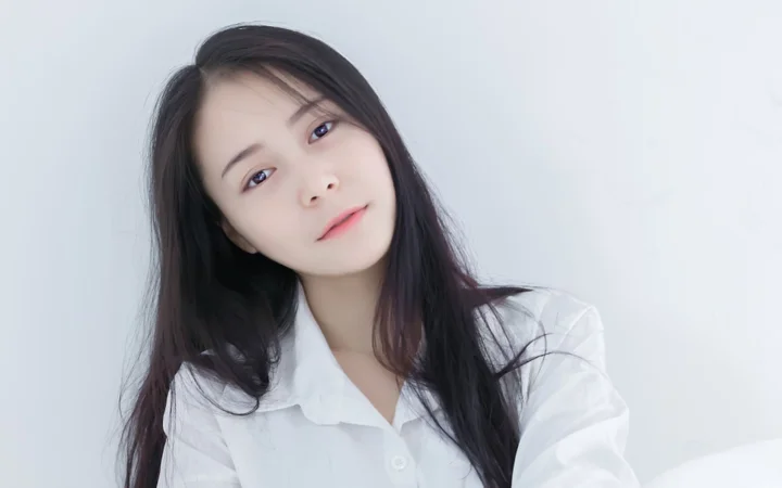 一位长发女性身穿白衬衫，在浅色背景前面向镜头。