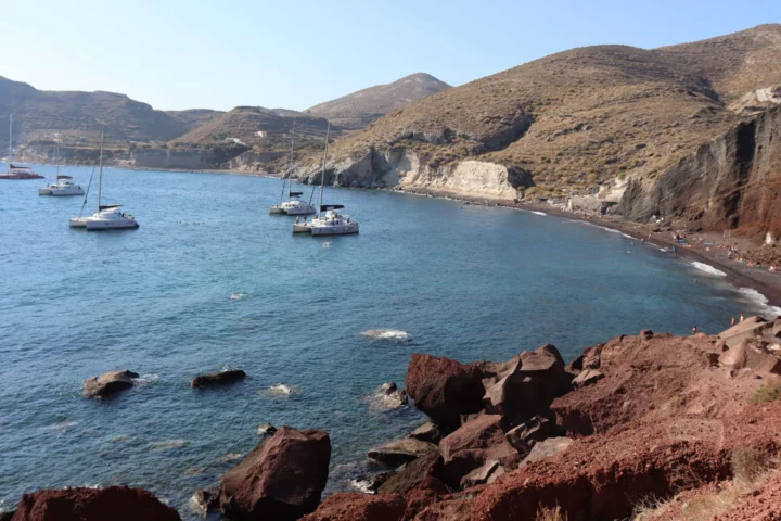 蓝色海湾中停泊着几艘帆船，岸边是红色岩石、陡峭山坡和狭长海滩。
