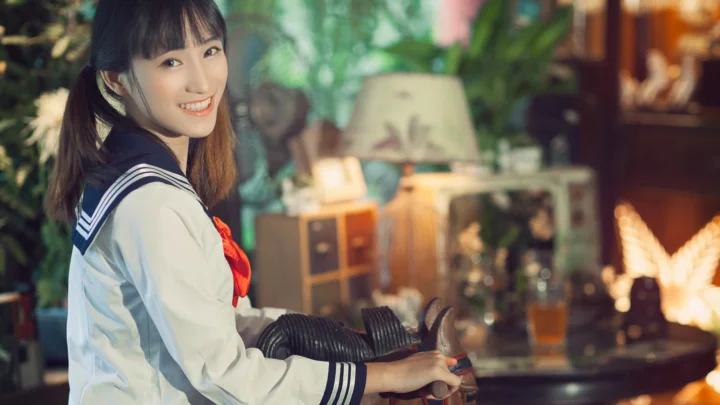 セーラー服風の衣装を着た女性が、室内でバイクのそばに立って微笑んでいます。