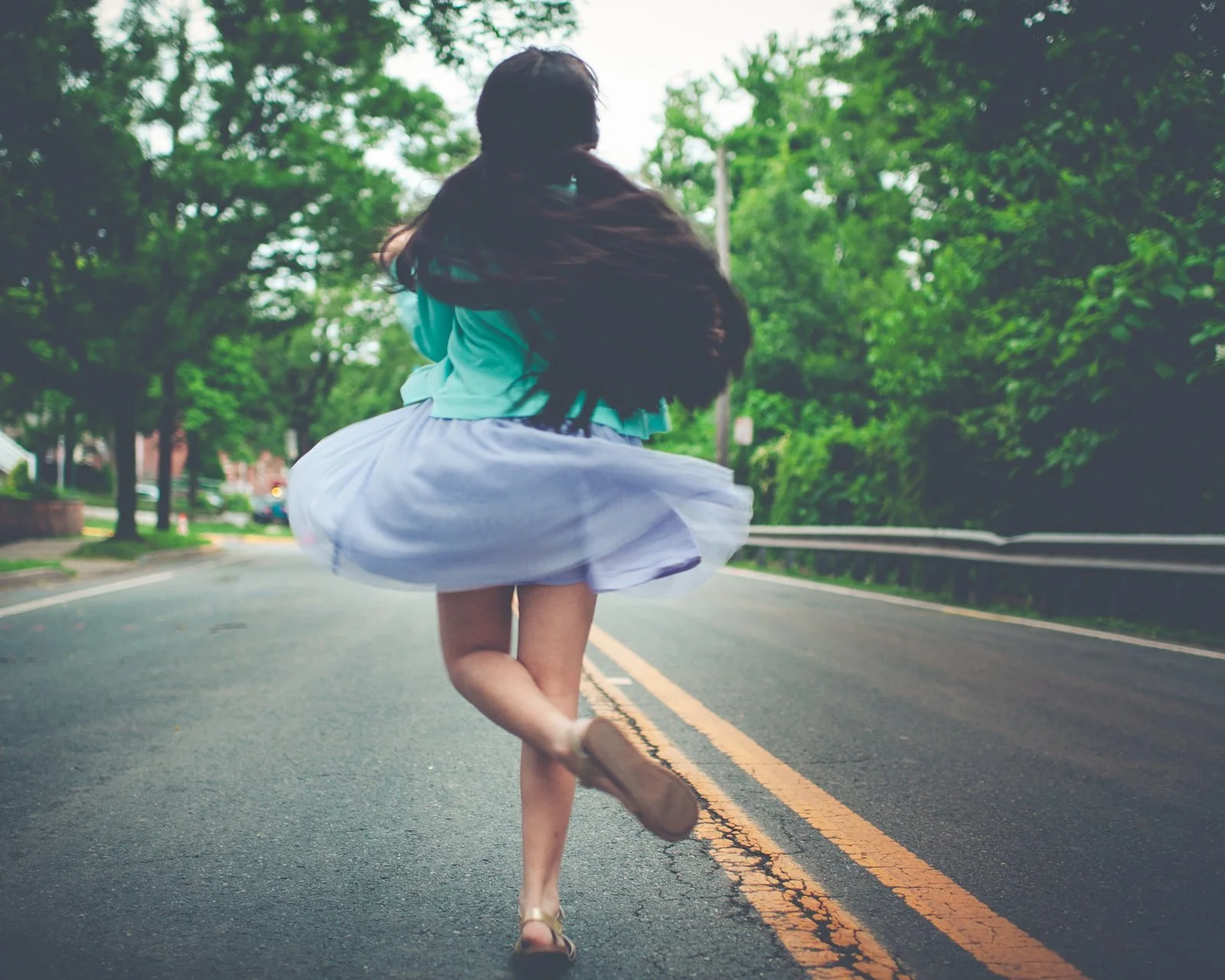 一名女子身穿青色上衣和白色裙子，在林荫道路上奔跑。