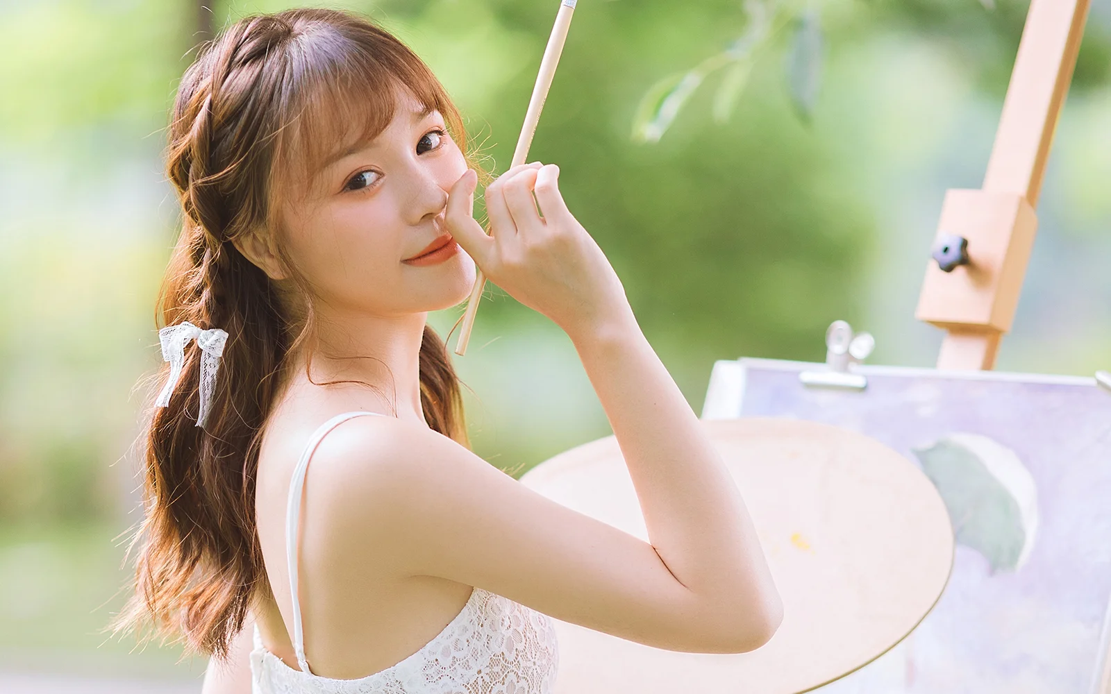 한 여성이 붓을 들고 야외 이젤 옆에 서 있으며 배경에는 부드러운 초록 풍경이 보입니다.