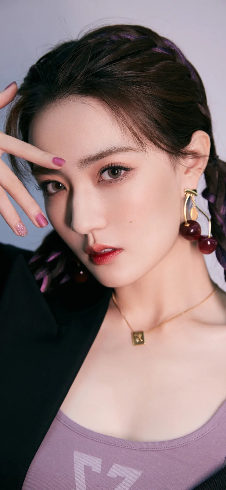 一位成年女性近距离面对镜头，戴着樱桃耳坠和项链，手指轻触脸侧，背景为浅紫色。