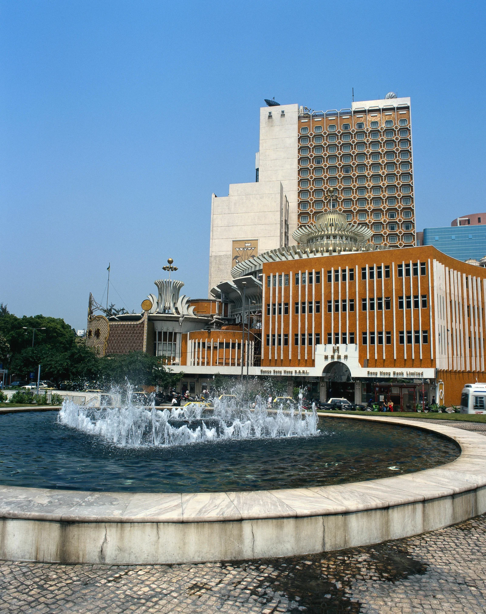 蓝天下，一座城市建筑矗立在喷泉水池后方。