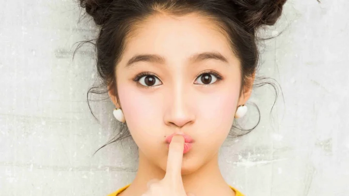 女性がカメラに向かい、明るい質感のある壁を背景に唇の前へ指を立てている。