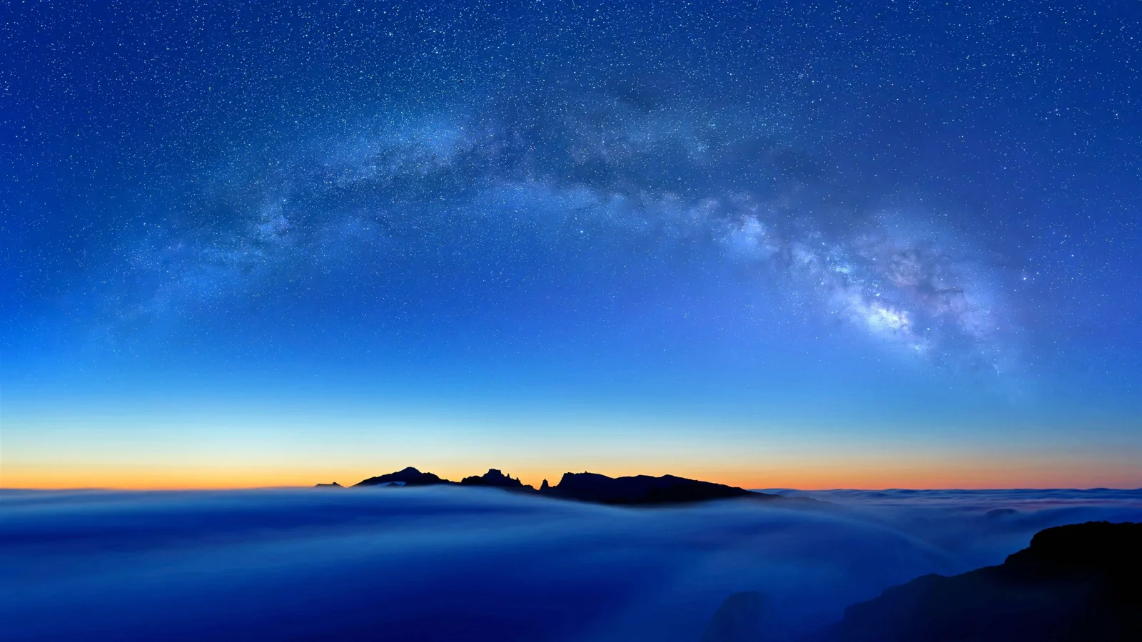 银河横跨深蓝色夜空，地平线处可见深色山脊和蓝色云海。