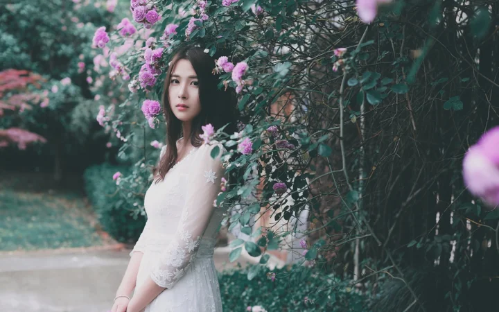 一名身穿白裙的女子站在开满粉色花朵的花园小径旁。