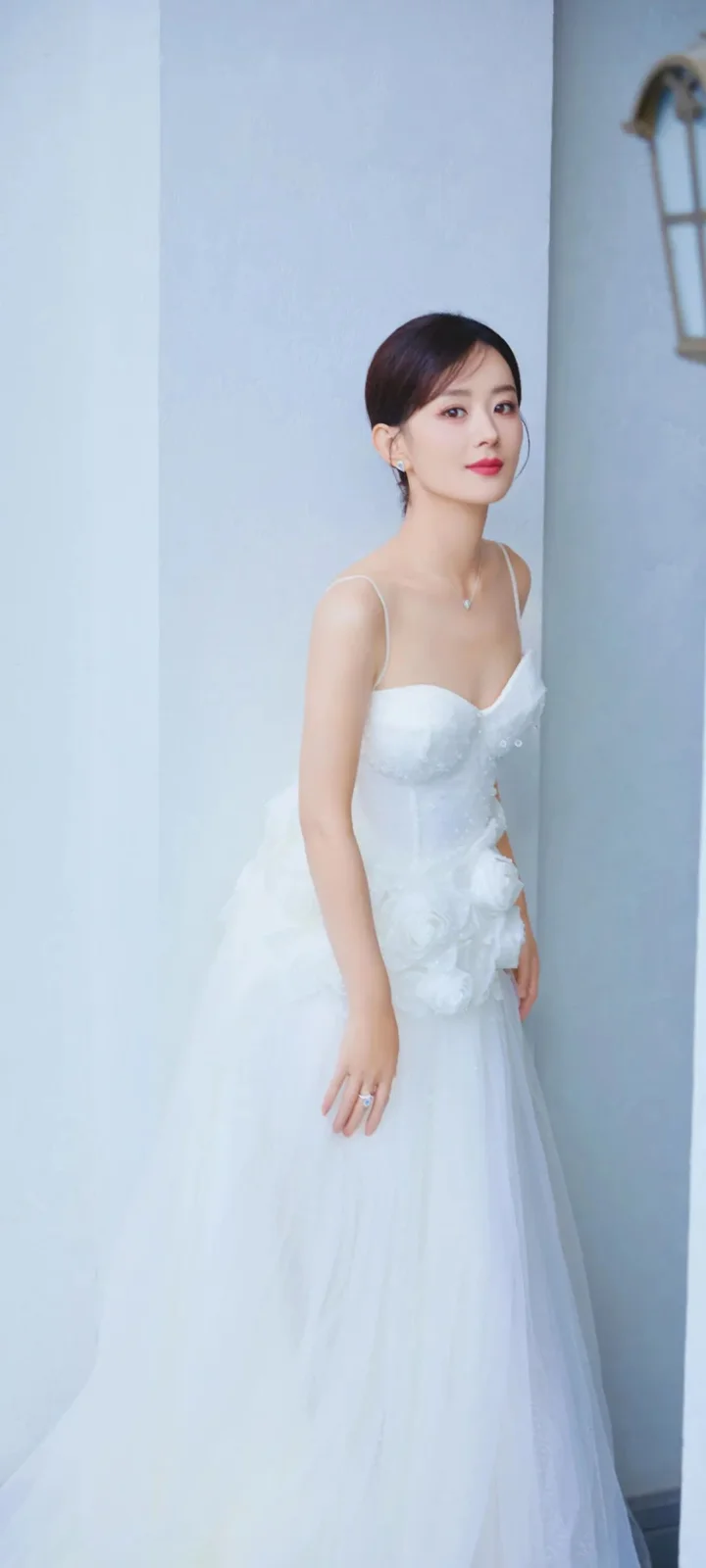 白いロングドレスを着た成人女性が淡い青色の室内背景の前に立っています。