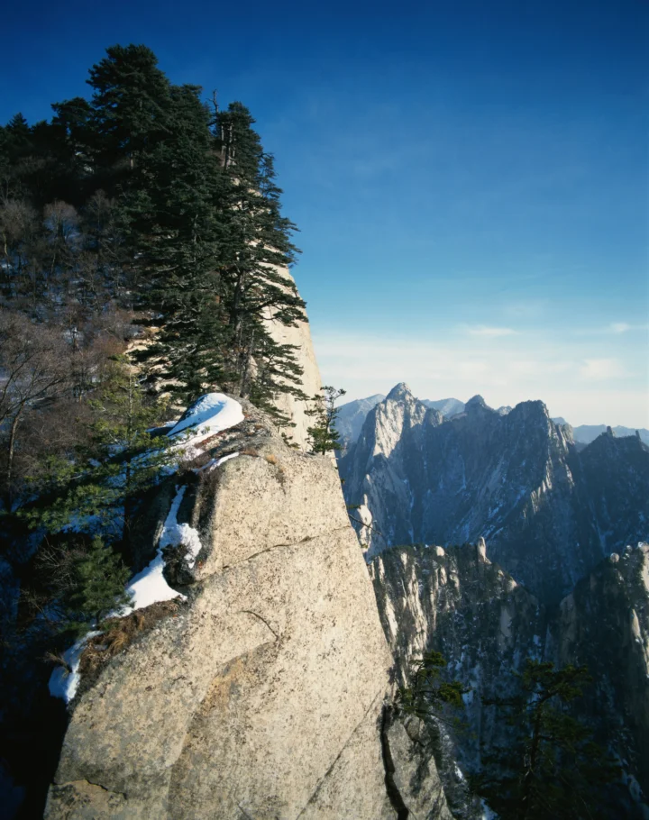 눈 덮인 바위 절벽과 침엽수림 너머로 먼 산맥이 펼쳐져 있습니다.