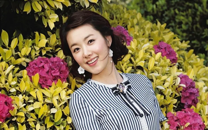 一名面带微笑的成年女子站在盛开花朵与绿叶环绕的户外环境中。