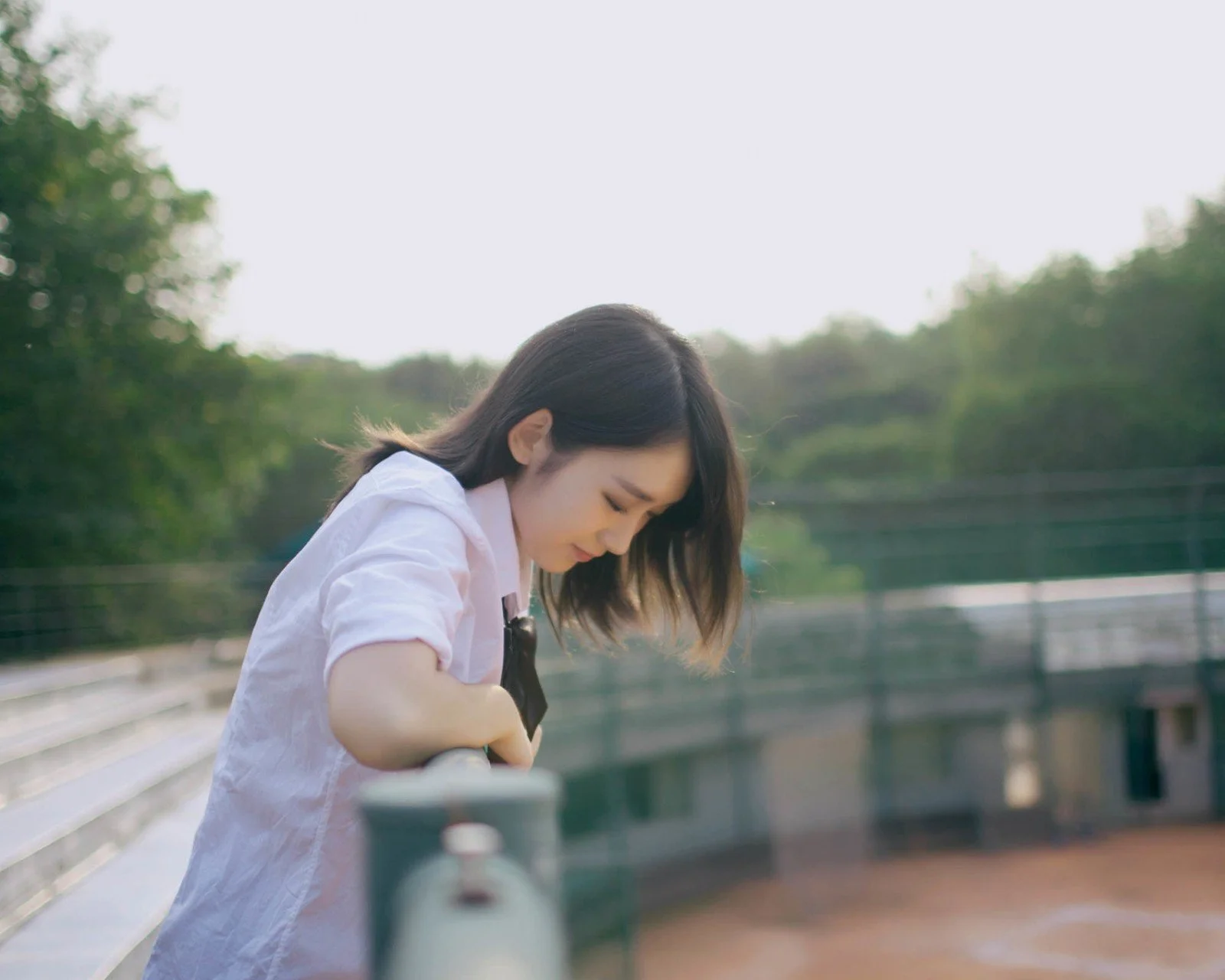 초록으로 둘러싸인 다리 옆에서 여성이 난간에 기대어 아래를 바라보고 있다.