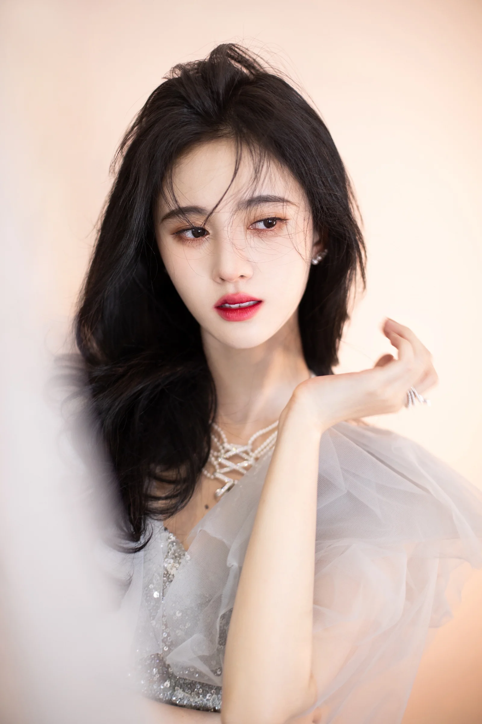 검은 머리의 여성이 밝은 색상의 시스루 드레스와 목걸이를 착용하고 부드러운 밝은 배경 앞에서 우아하게 포즈를 취하고 있습니다.