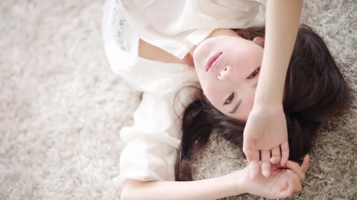 一名身穿白色衣服的女子躺在柔软地毯上，手臂举过头顶看向镜头。