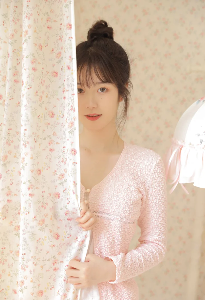 ピンクのドレスを着た女性が、柔らかな室内光の中で花柄のカーテンのそばに立っています。