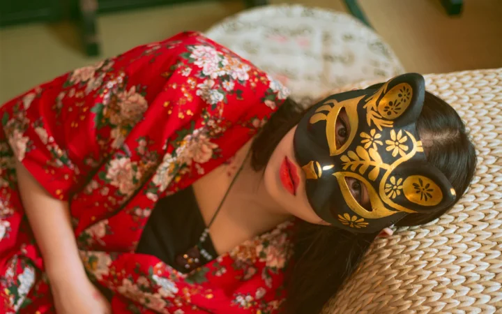 一名戴着黑金面具、身穿红色花卉连衣裙的成年女子侧躺在编织软垫上。