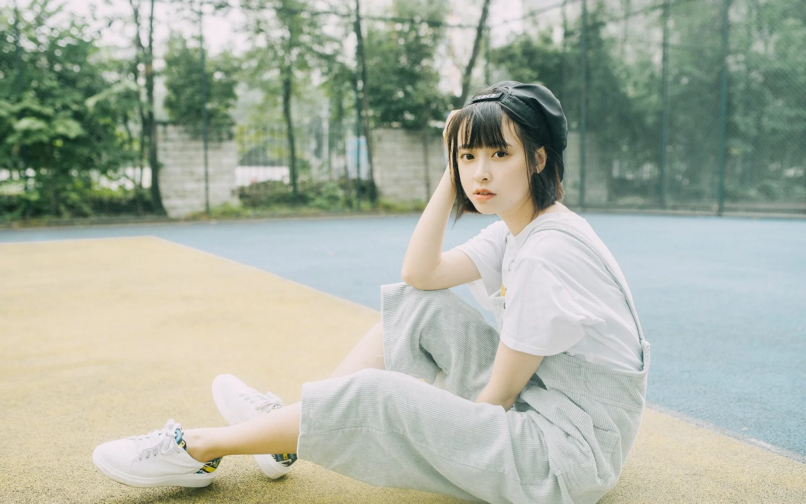 一名戴黑色帽子的年轻女性坐在户外球场上，身后是树木和围栏。