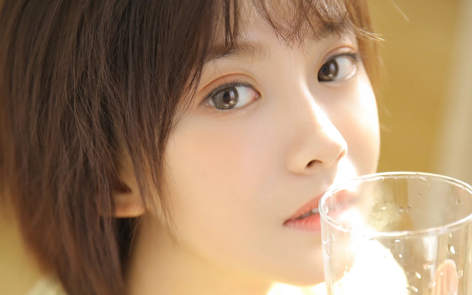一位女性在暖色光线下手持透明玻璃杯，面部特写占据画面大部分。