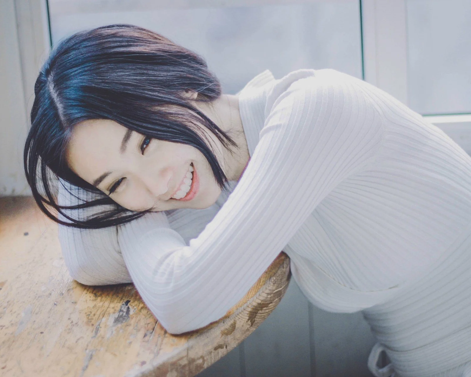白い長袖の服を着た女性が木のテーブルに寄りかかって微笑んでいる。