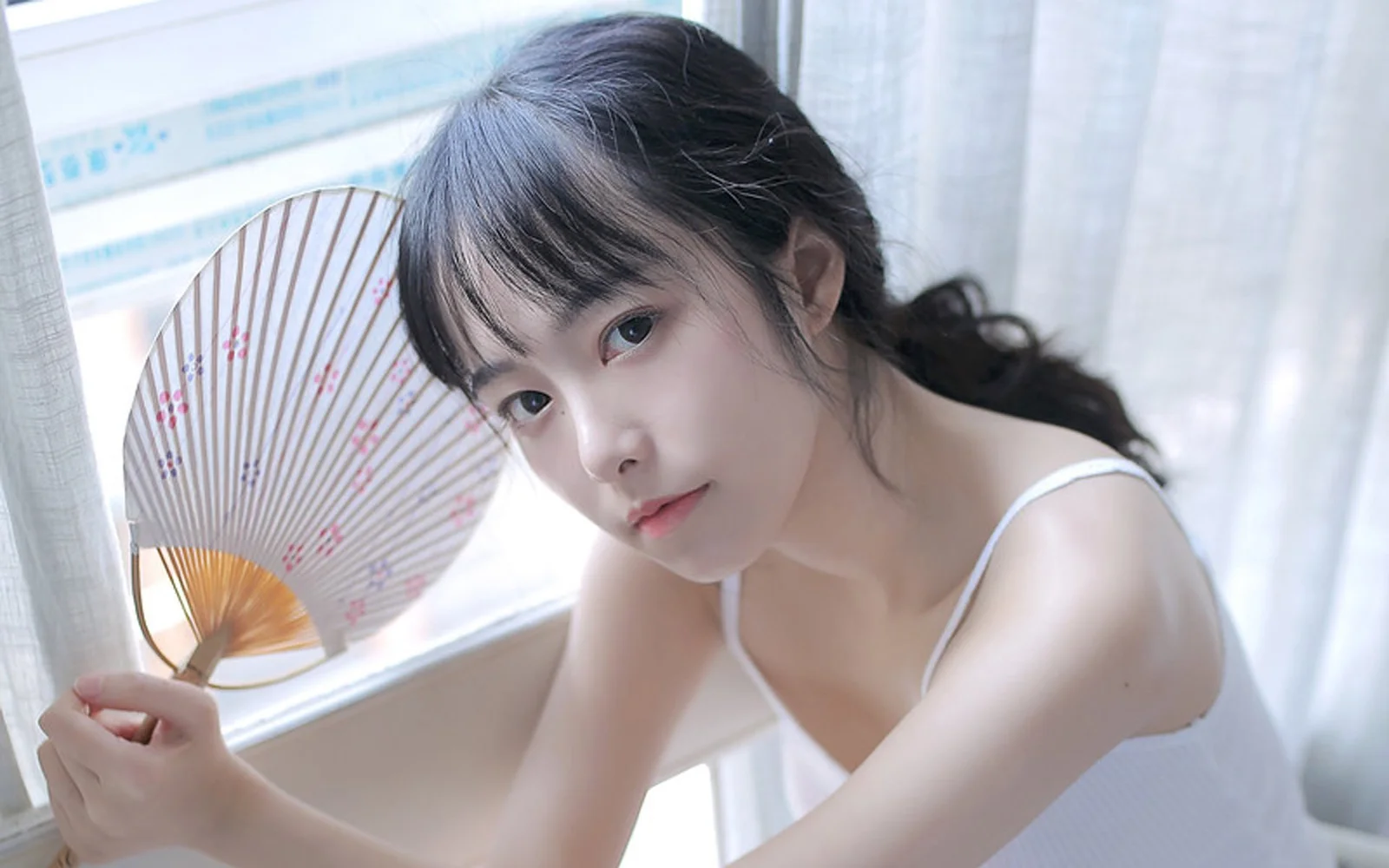一名女子在窗边手持折扇，柔和的室内光线映照在她身上。