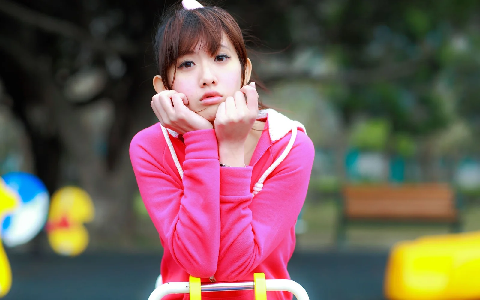 분홍색 후드티를 입은 여성이 야외에서 두 손으로 턱을 괴고 카메라를 바라보고 있습니다.
