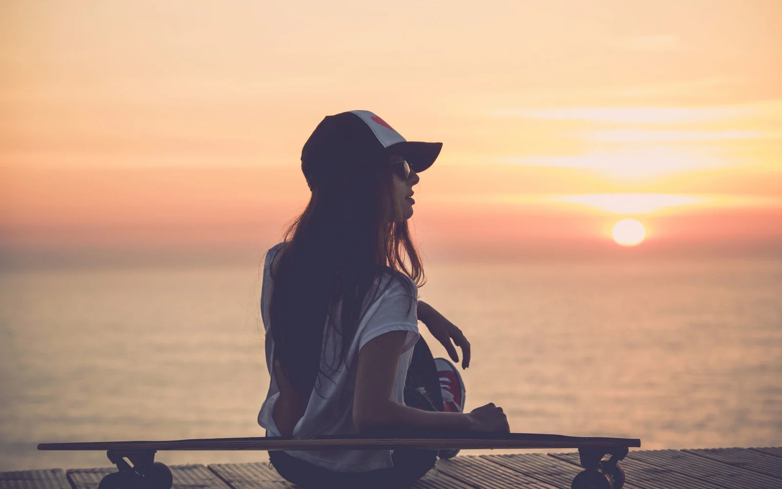 一名戴帽子的女性坐在海边木平台上，夕阳映照着平静的海面。