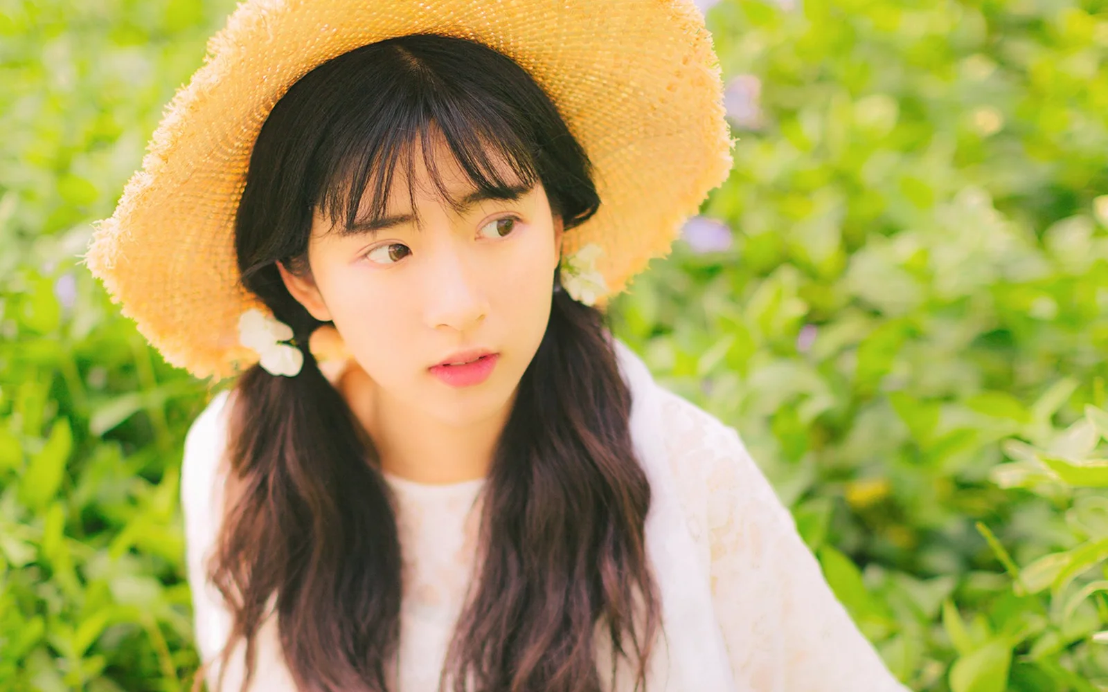 麦わら帽子をかぶった若い女性が、緑の葉を背景に横を見ている。