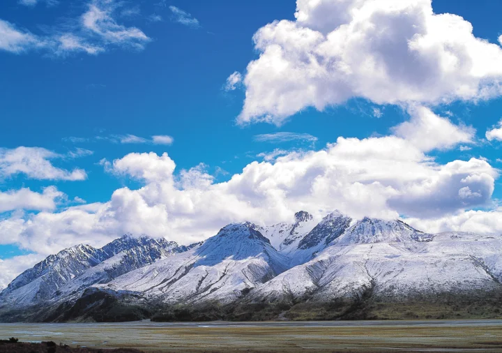 积雪覆盖的群山矗立在蓝天白云下，前景是一片平缓的河岸或湿地。