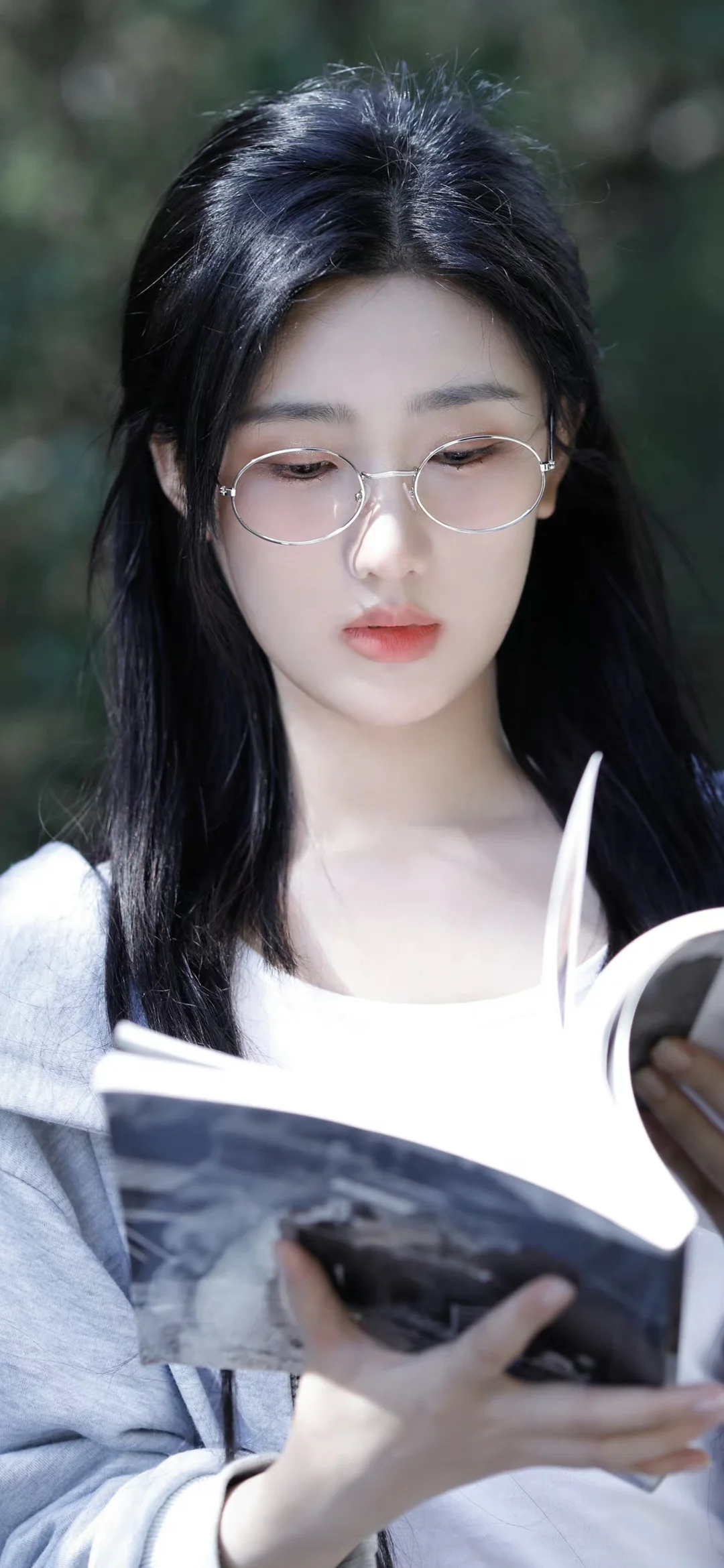 一名戴圆框眼镜的女子手持书本站在虚化的绿色背景前。