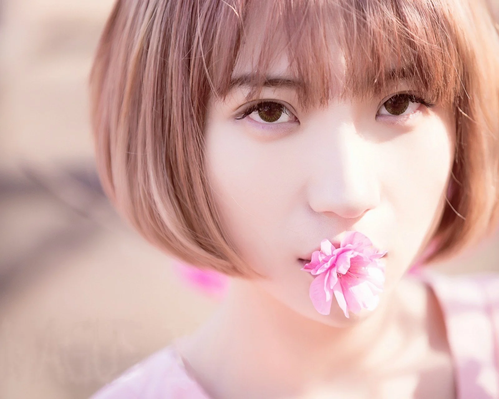 短い髪の女性が唇元にピンク色の花を添えたクローズアップポートレートです。