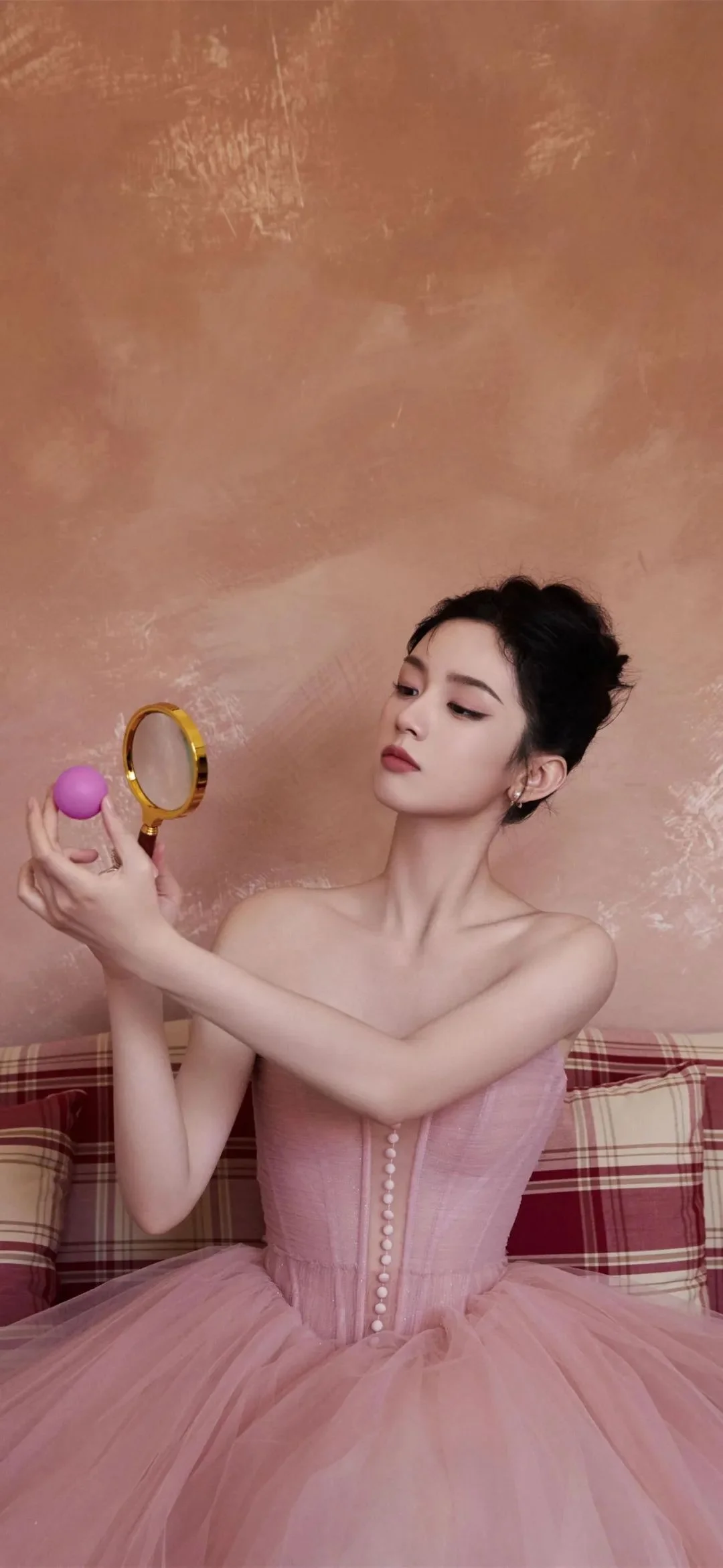 一名成年女性身穿粉色礼服，手持小镜子和紫色圆球坐在格纹沙发前。