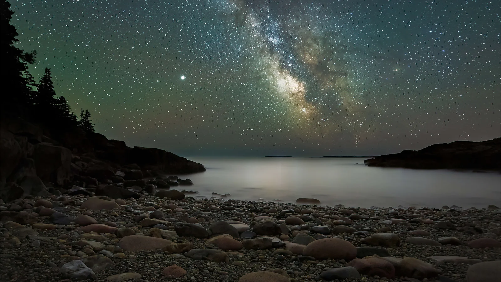 银河横跨繁星密布的夜空，下方是宁静海面与布满圆石的海岸。