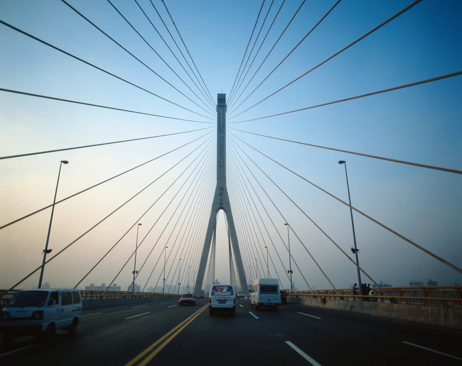 一座悬索桥横跨城市道路，缆索从桥塔向四周延伸，车辆行驶在桥面上。