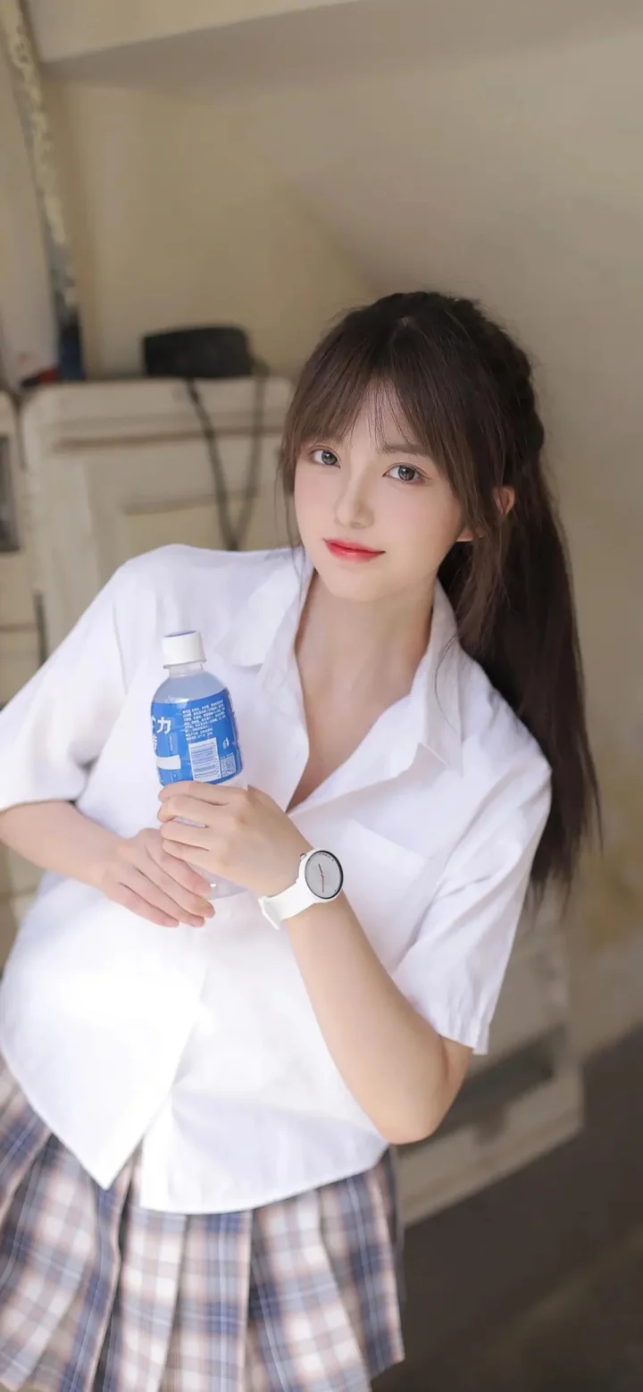 一名长发女生穿着白衬衫，手持一瓶水站在室内。