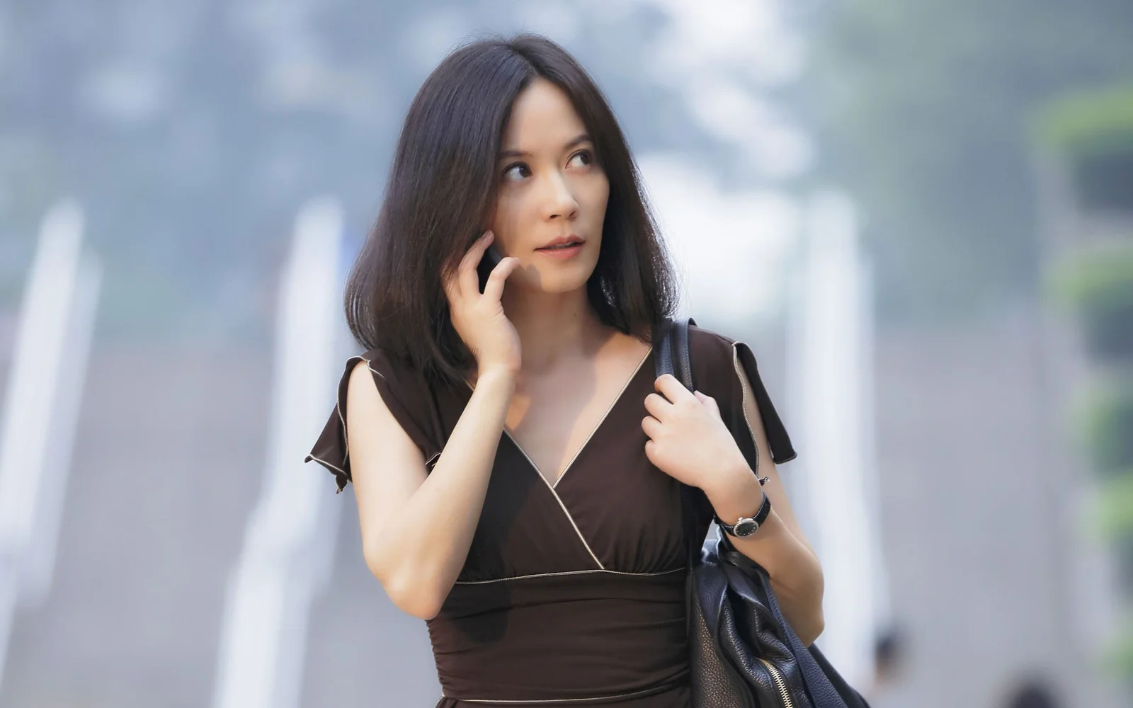 어두운 원피스를 입은 여성이 흐릿한 도시 배경 앞에서 검은색 핸드백을 들고 서 있습니다.