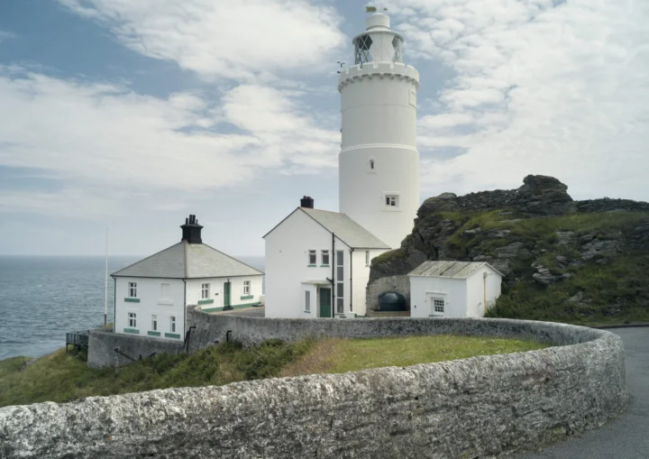 一座白色灯塔和附属建筑坐落在英国岩石海岸边，远处是海洋与多云天空。