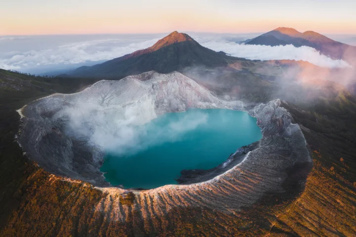 一座 turquoise 色火山口湖 surrounded by rugged volcanic ridges and distant mountains above the clouds.