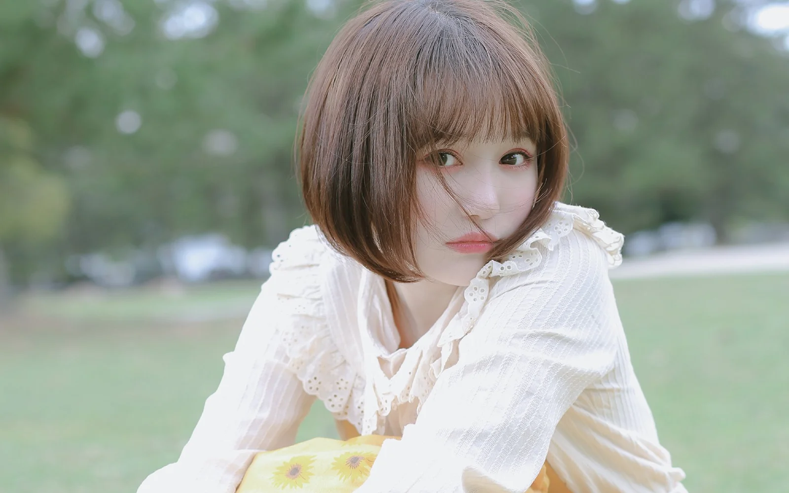 白いブラウスを着たショートヘアの女性が、屋外の芝生を背景にカメラを見ています。