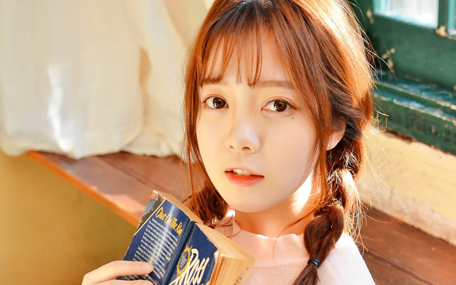 갈색 긴 머리의 여성이 실내 햇빛 아래에서 펼친 책을 들고 카메라를 바라보고 있다.