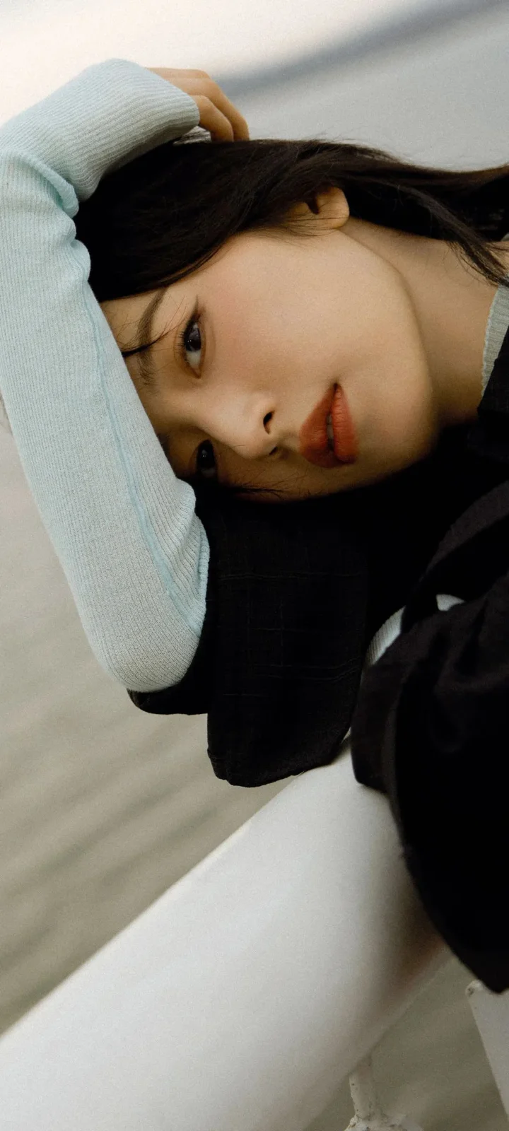 一名黑发女性侧卧在浅色背景上，手臂搭在头顶附近，面部占据画面大部分。