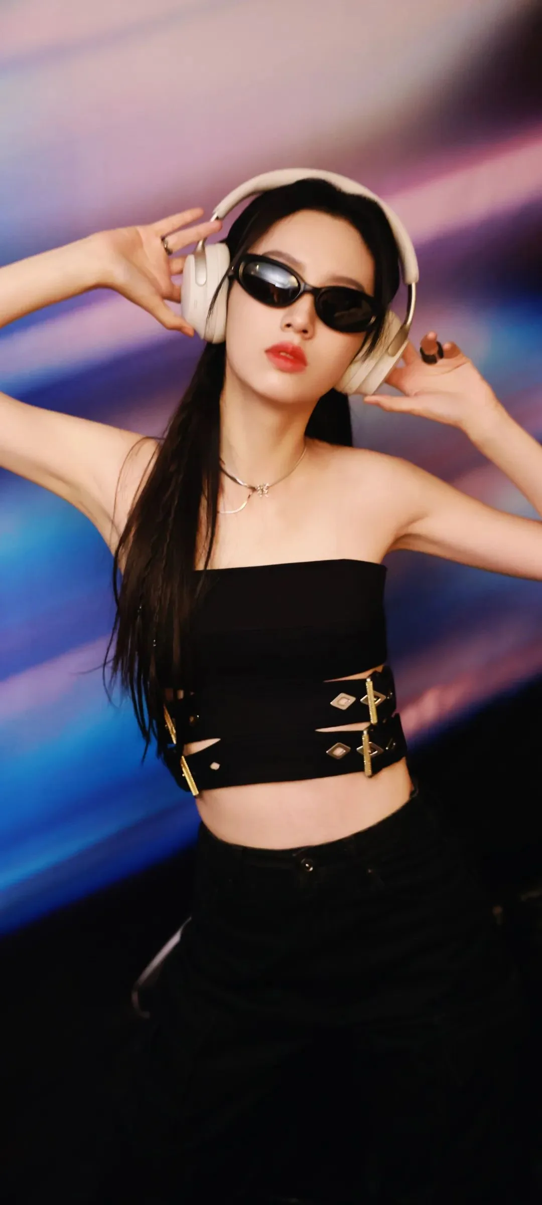 一名戴着耳机和墨镜的女性站在彩色光影背景前摆姿势。