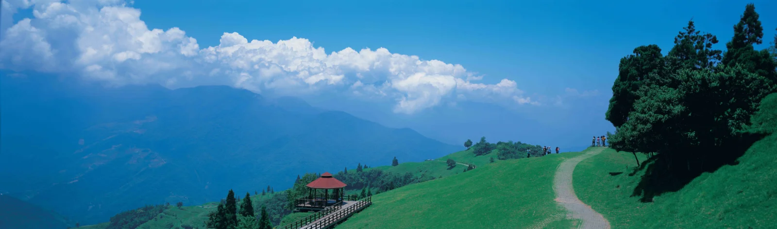 綠色山坡上的小徑通往遠處的層疊群山，藍天中飄著大片白雲。