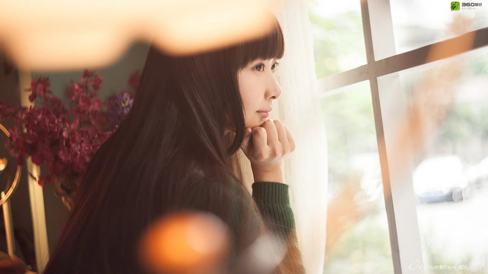 一名女子坐在窗边，侧脸望向窗外，前景带有柔和的虚化光斑。