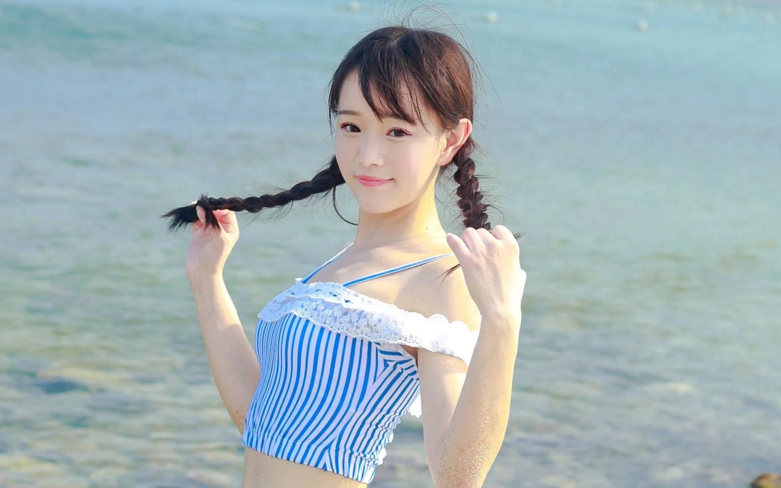 양갈래 땋은 머리의 여성이 얕은 바닷물가에서 머리를 정리하고 있다.